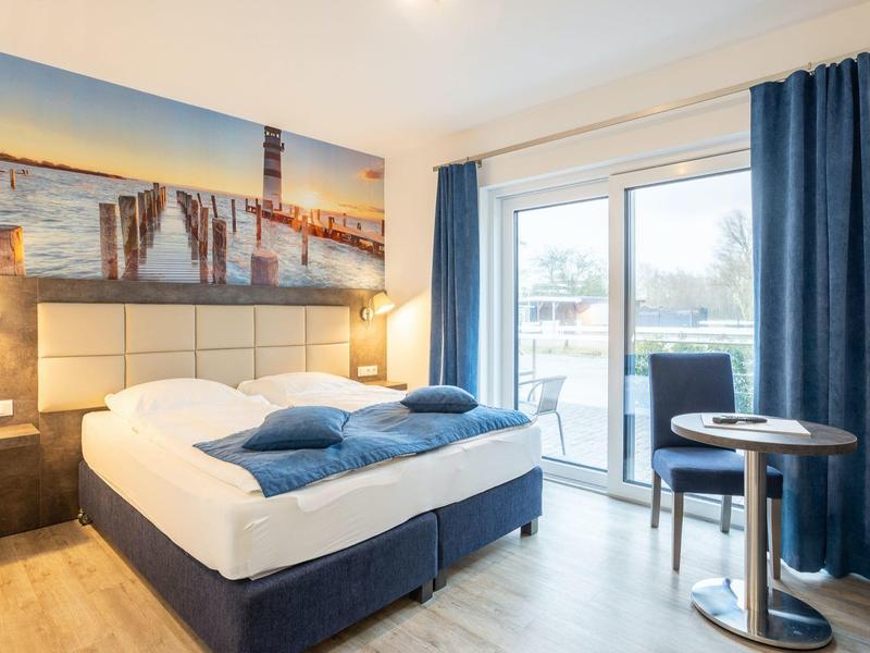 Helles Schlafzimmer mit Doppelbett, blauen Vorhängen, Balkon und maritimer Wanddekoration.