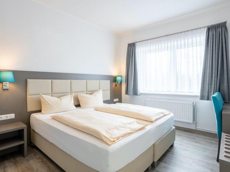 Helles Schlafzimmer mit Doppelbett, weißen Wänden, grauem Vorhang und blauem Schreibtischstuhl.