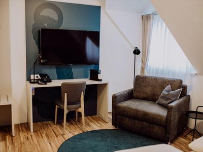 Pohodlný hotelový pokoj s TV, pracovním stolem, křeslem a přirozeným světlem.