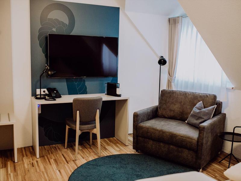 Pohodlný hotelový pokoj s TV, pracovním stolem, křeslem a přirozeným světlem.