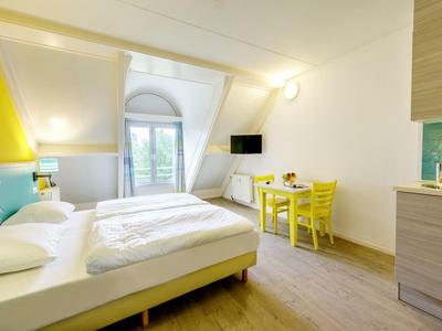 Lichte hotelkamer met tweepersoonsbed, gele stoelen, bureau en wand-tv onder een dakraam.