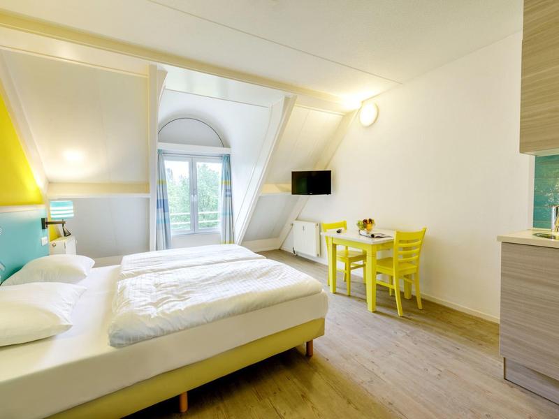 Lichte hotelkamer met tweepersoonsbed, gele stoelen, bureau en wand-tv onder een dakraam.