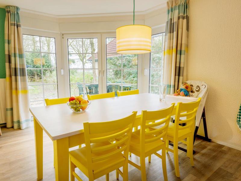 Eetkamer met gele tafel en stoelen, grote ramen en uitzicht op de tuin.