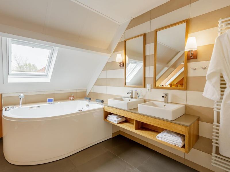 Lichte badkamer met hoekbad, twee wastafels en grote spiegels onder een schuine dak.
