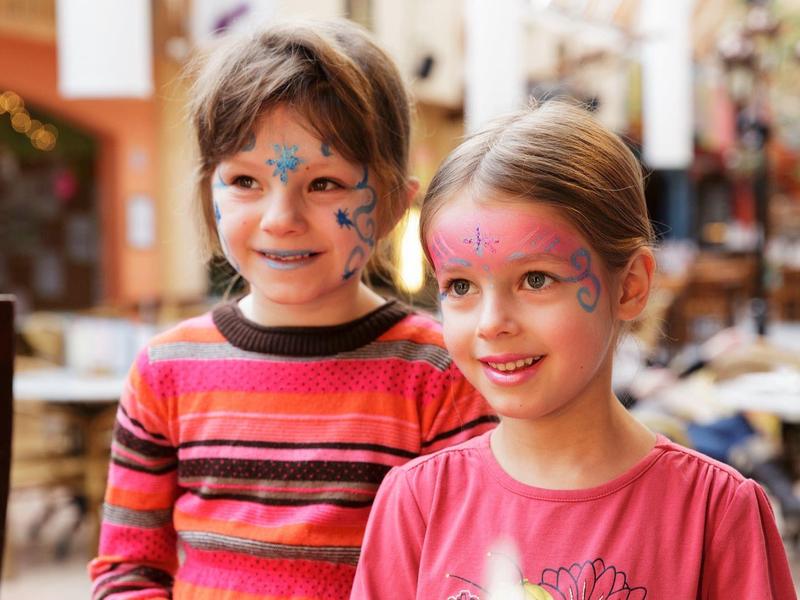 Twee kinderen met geschilderde gezichten glimlachen in een gezellige binnenruimte.