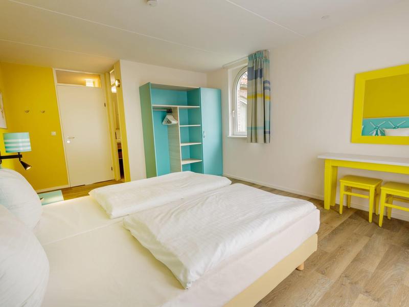 Lichte hotelkamer met gele accenten, een bed, bureau en open kast.