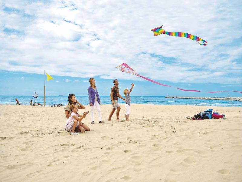 Des gens font voler des cerfs-volants sur une plage ensoleillée avec un ciel partiellement nuageux.