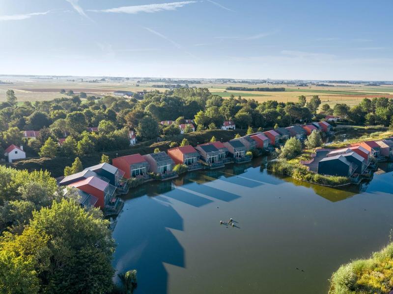 Domki letniskowe nad brzegiem spokojnego jeziora z zielonym krajobrazem i szerokim niebem w tle.