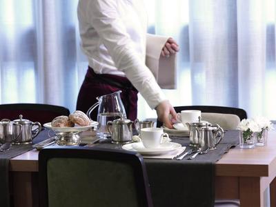 Ein gedeckter Frühstückstisch mit Tee- und Kaffeekannen, Tassen und Brötchen, Frau im Hintergrund.