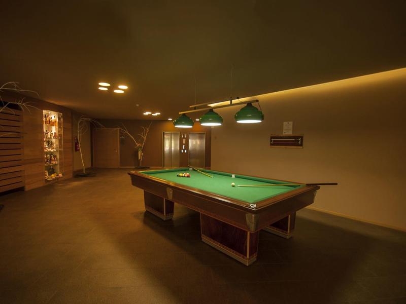 Ein großer, gedämpft beleuchteter Billardraum mit grünem Tisch und Treppe im Hintergrund.