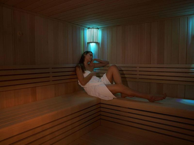 Frau mit Handtuch sitzt entspannt auf Holzbank in gedämpft beleuchteter Sauna.