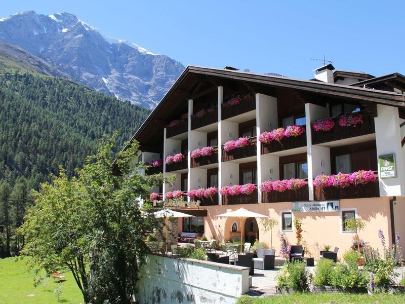 Großes Hotel mit Balkonen und pinken Blumen, umgeben von grünen Wiesen und Bergen im Hintergrund.