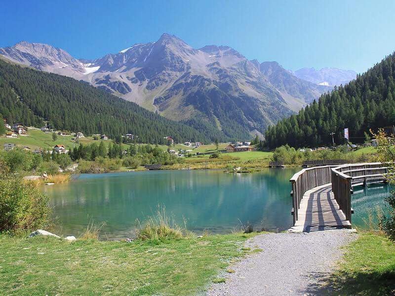 Kleiner Bergsee mit grünem Wasser, Umgeben von Wiesen, Nadelwäldern und hohen Bergen im Hintergrund.