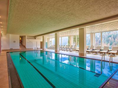 Innenbereich eines Schwimmbads mit klarem Wasser, Liegestühlen und großen Fenstern.