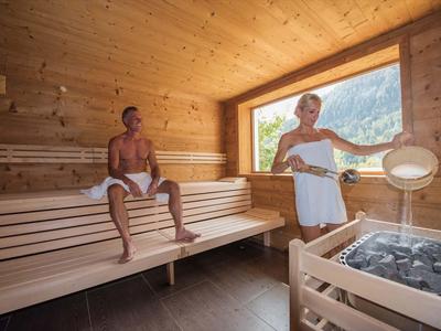 Zwei Personen entspannen in einer Holzsauna mit großer Fensterfront und Blick ins Grüne.