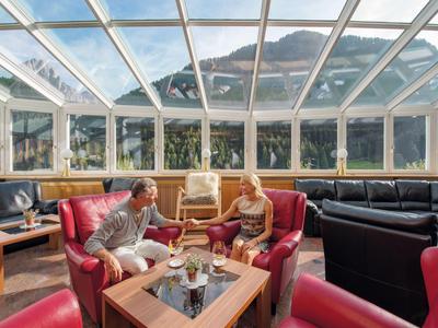 Zwei Personen sitzen in einem hellen Wintergarten mit Bergblick und roten Sesseln.