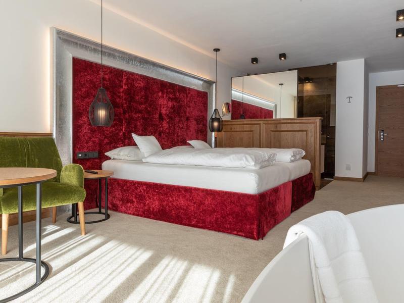 Modernes Schlafzimmer mit rotem Velourbett, grünem Stuhl und hellem Holzmöbel im Tageslicht.