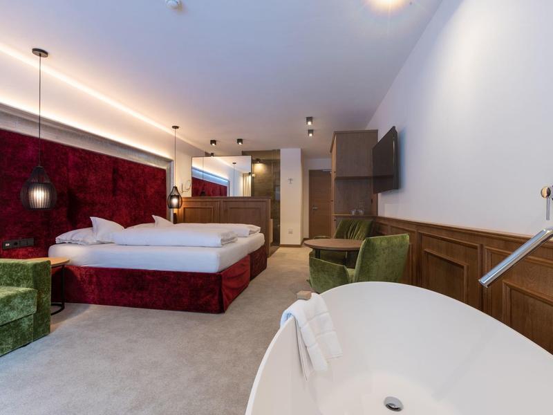 Modernes Hotelzimmer mit Doppelbett, rotem Kopfteil, grünem Sessel und freistehender Badewanne.
