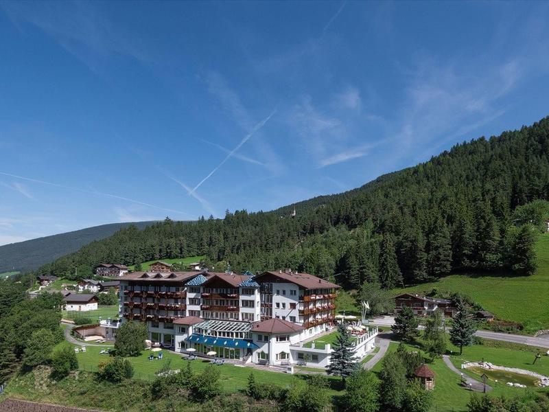 Berghotel mit Terrassenpool, umgeben von grünen Wiesen und dichten Nadelwäldern unter blauem Himmel.