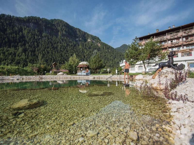 Klare Bergquelle mit Felsen, grünen Wäldern und einem großen Gebäude am Ufer unter blauem Himmel.