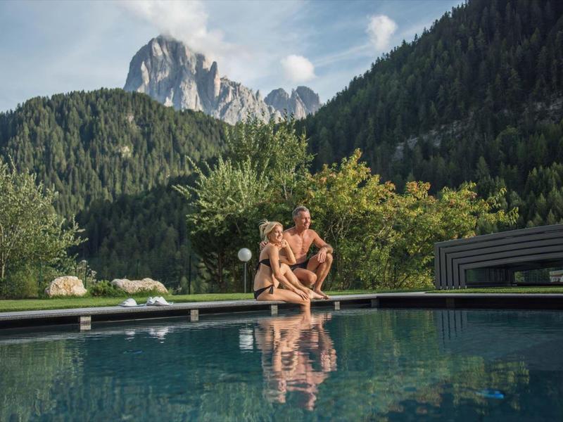 Mann sitzt entspannt am Rand eines Pools vor bewaldeten Bergen und blauem Himmel.
