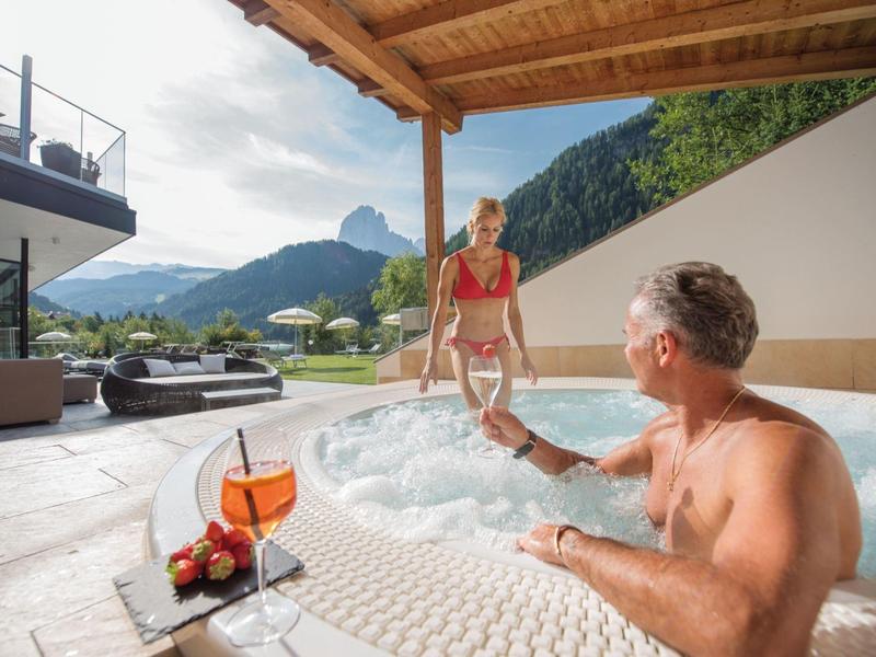 Mann und Frau im Whirlpool mit Bergblick, Terrasse mit Loungemöbeln und Snackplatte nebenan.