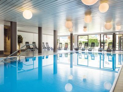 Hell beleuchteter Hallenbadbereich mit blauer Wasseroberfläche und Liegestühlen am Rand.