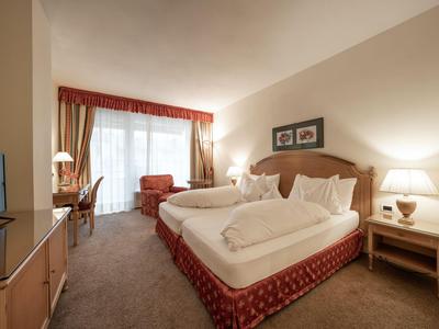 Gemütliches Hotelzimmer mit Doppelbett, Sessel, Schreibtisch und Balkon bei Tageslicht.