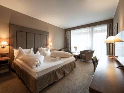 Modernes Hotelzimmer mit großem Bett, Stühlen am Fenster und Fernseher an der Wand.