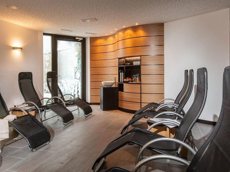 Ruheraum mit schwarzen Liegestühlen, Holzwand, sanfter Beleuchtung und Glastür zum Außenbereich.