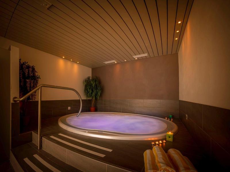 Runder Whirlpool mit violettem Wasser in warm beleuchtetem Spa-Raum mit Kerzen und Pflanzen.