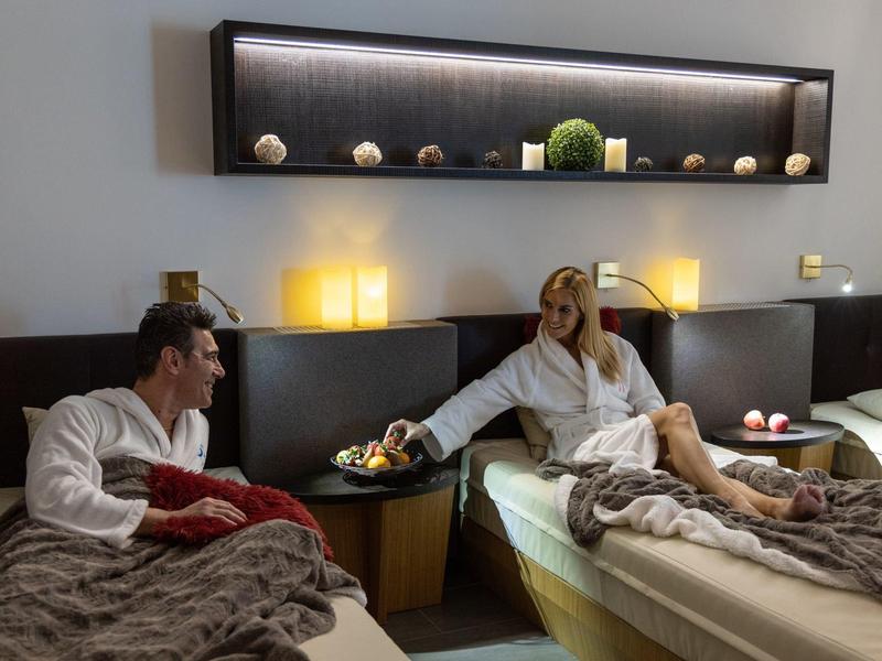 Zwei Personen in weißen Bademänteln sitzen auf Betten in einem warm beleuchteten, modernen Hotelzimmer.
