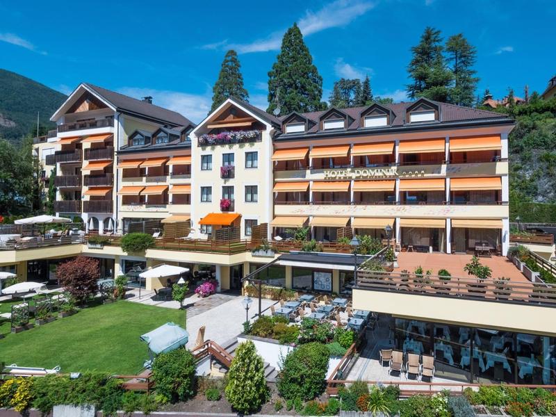 Großes alpines Hotel mit Holzverkleidung, Terrasse, grüner Wiese und Bergen im Hintergrund.