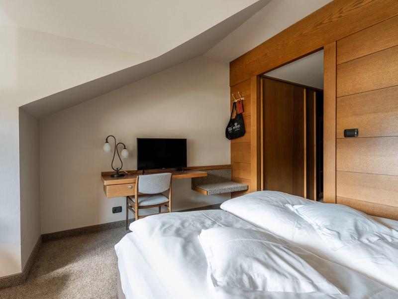 Helles Schlafzimmer mit Dachschräge, weißer Bettdecke, Holztür und Schreibtisch mit Stuhl und Fernseher.