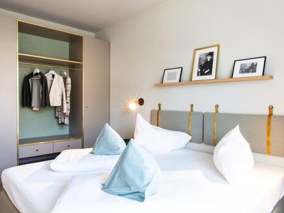 Weißes Doppelbett mit blauen Kissen, Holzwandregal mit Bildern, offener Kleiderschrank mit Kleidung.