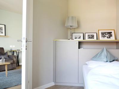 Kleines Schlafzimmer mit weißer Wand, Einzelbett, Regal und grauer Couch im Hintergrund.