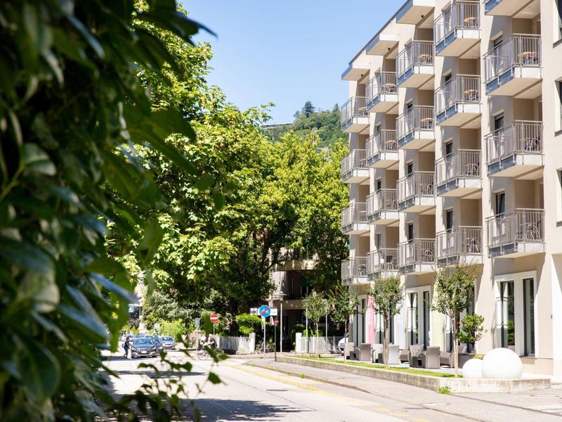 Straße mit mehrstöckigem Wohnhaus, grünen Bäumen und hellblauem Himmel im Hintergrund.