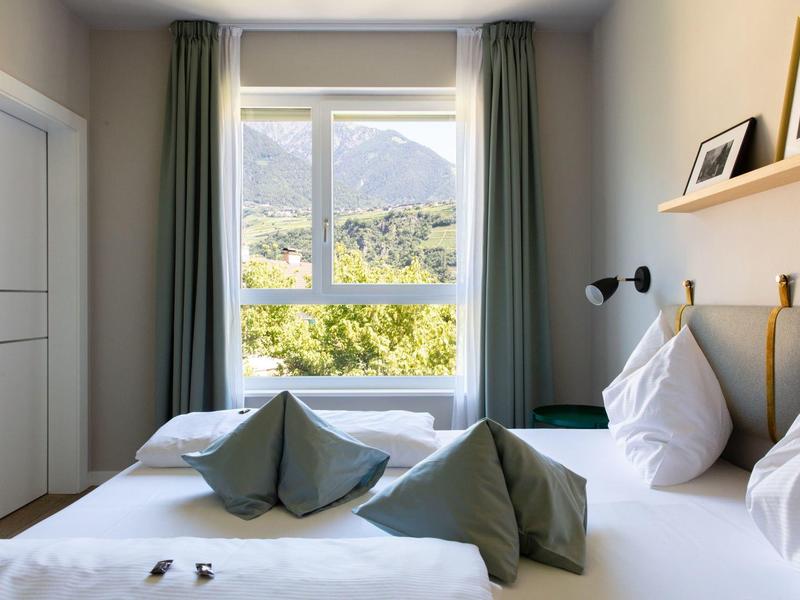 Hell eingerichtetes Schlafzimmer mit großem Fenster, grünen Vorhängen und Bergblick draußen.