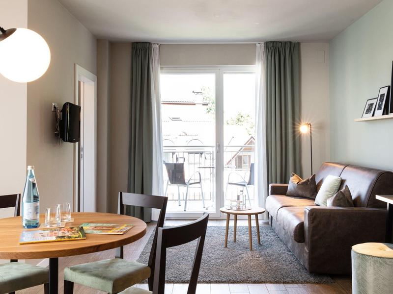 Helles Wohnzimmer mit grauem Sofa, runden Holztisch und Balkon hinter großen Fenstern.