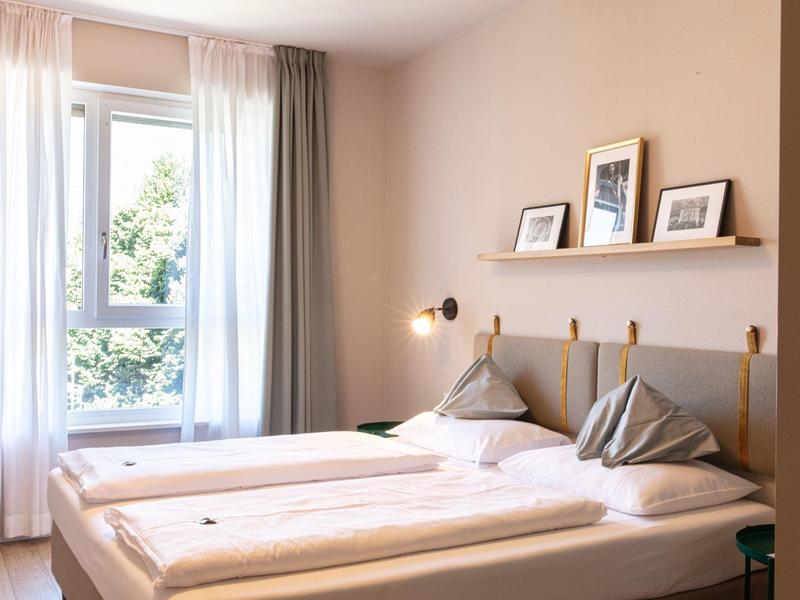 Helles Schlafzimmer mit zwei Einzelbetten, grauen Kissen, weißen Vorhängen und drei gerahmten Bildern.