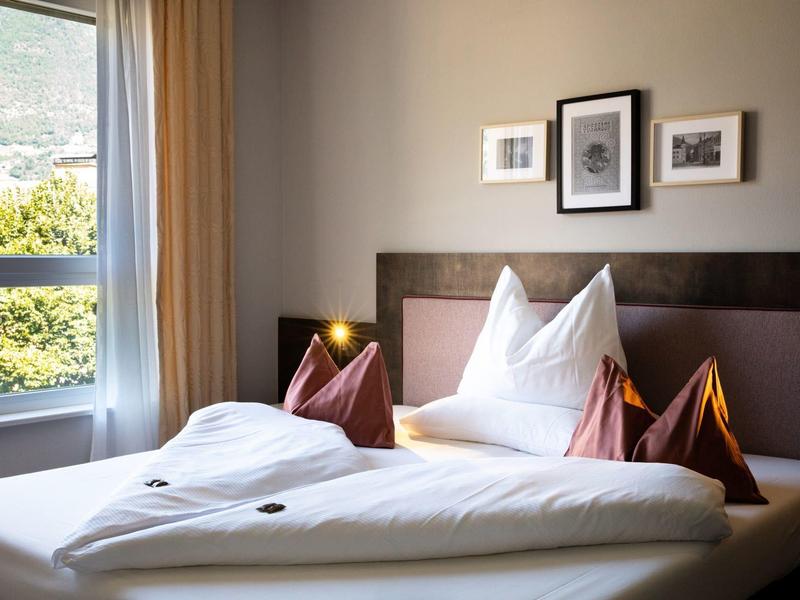 Gemütliches Schlafzimmer mit großem Fenster, Doppelbett, weißen und braunen Kissen, drei Bildern an der Wand.