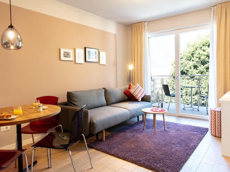 Helles Wohnzimmer mit grauem Sofa, rundem Holztisch, lila Teppich und Balkon mit grünen Bäumen.