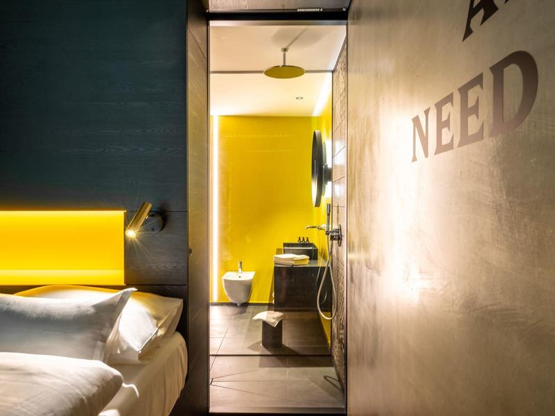 Modernes Hotelzimmer mit gemütlichem Bett und Blick ins Bad mit gelber Akzentbeleuchtung.