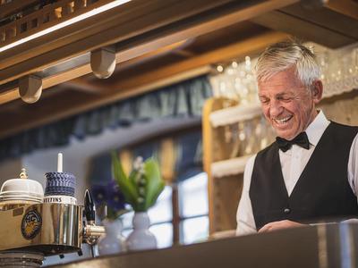 Älterer Kellner in schwarzer Weste lächelt hinter rustikalem Barbereich mit Holzdecke und Glasregal.
