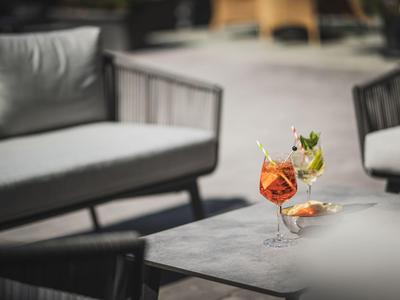 Zwei bunte Cocktails stehen auf einem grauen Tisch vor modernen, grauen Sitzmöbeln im Freien.