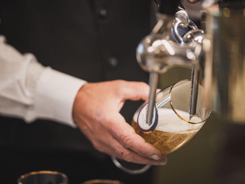 Eine Hand hält ein Glas schräg unter einen Zapfhahn, Bier mit Schaum fließt hinein.