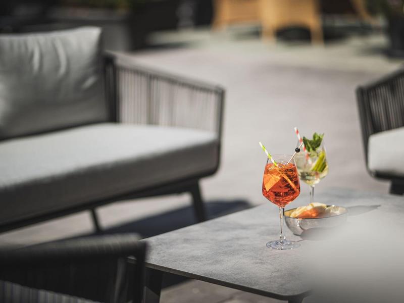 Zwei bunte Cocktails stehen auf einem grauen Tisch vor modernen, grauen Sitzmöbeln im Freien.