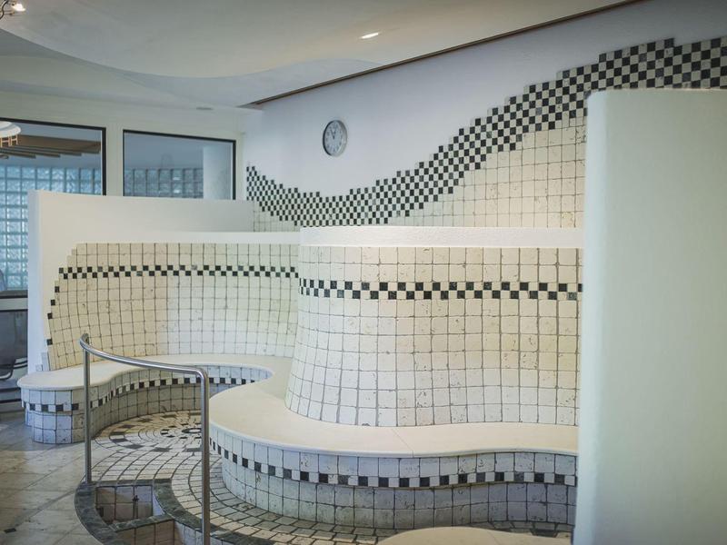 Moderner Wellnessbereich mit geschwungenen, mosaikverzierten Sitzbänken und großen Fenstern.