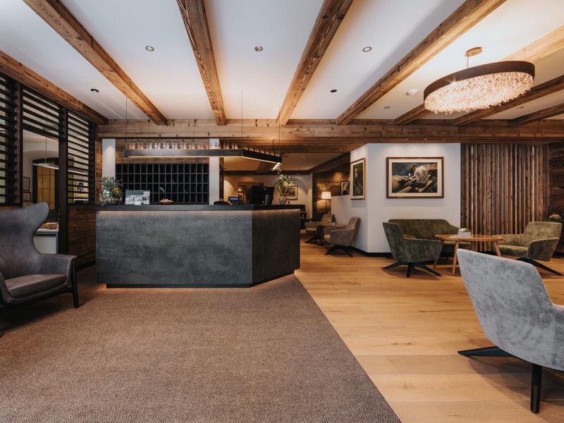 Moderner Hotel-Empfang mit Holzbalken, grauem Tresen, Sesseln und warmem Licht.