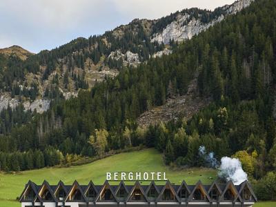 Mehrstöckiges Berghotel vor bewaldetem Berg mit wolkigem Himmel.
